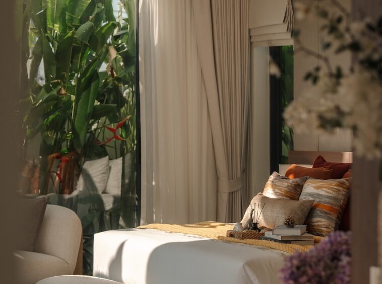 Luxurious bedroom with a white bed, cushioned bench, and warm lighting; large windows reveal tropical plants outside.
