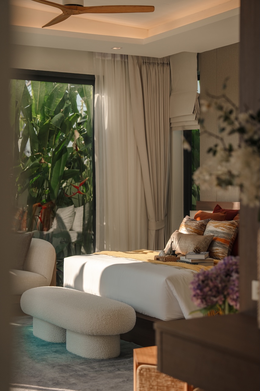 Luxurious bedroom with a white bed, cushioned bench, and warm lighting; large windows reveal tropical plants outside.