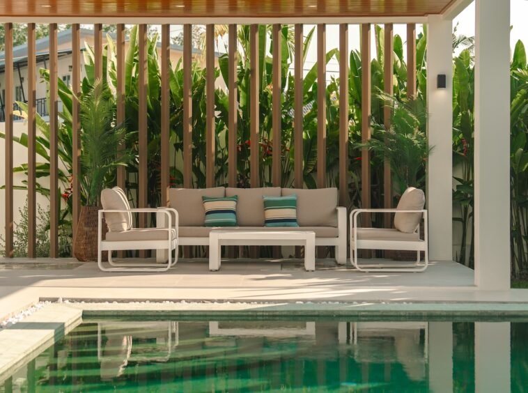 Outdoor poolside lounge with beige cushioned sofa and chairs, white frames, and tropical plants behind a wooden slat screen.