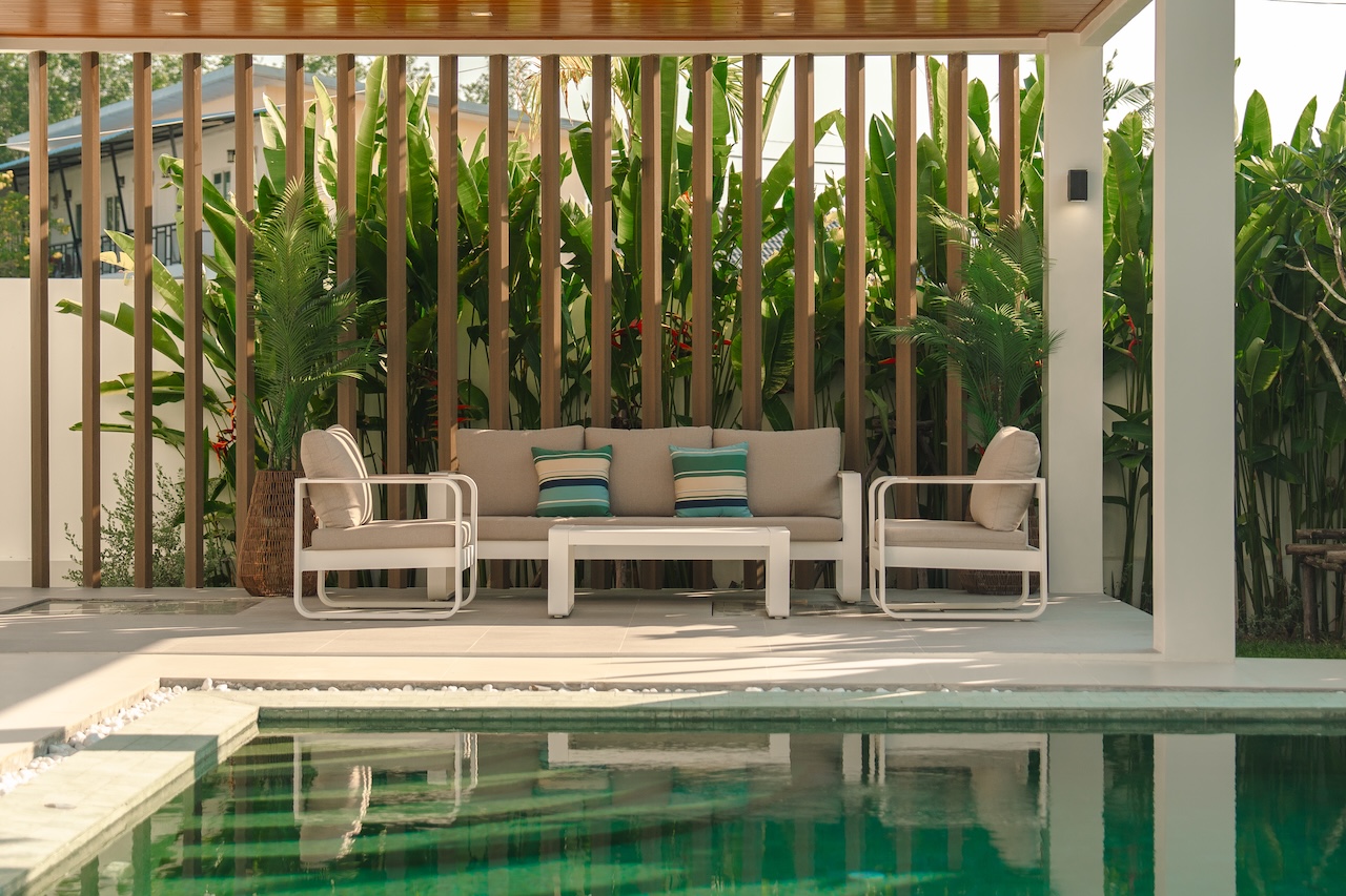 Outdoor poolside lounge with beige cushioned sofa and chairs, white frames, and tropical plants behind a wooden slat screen.