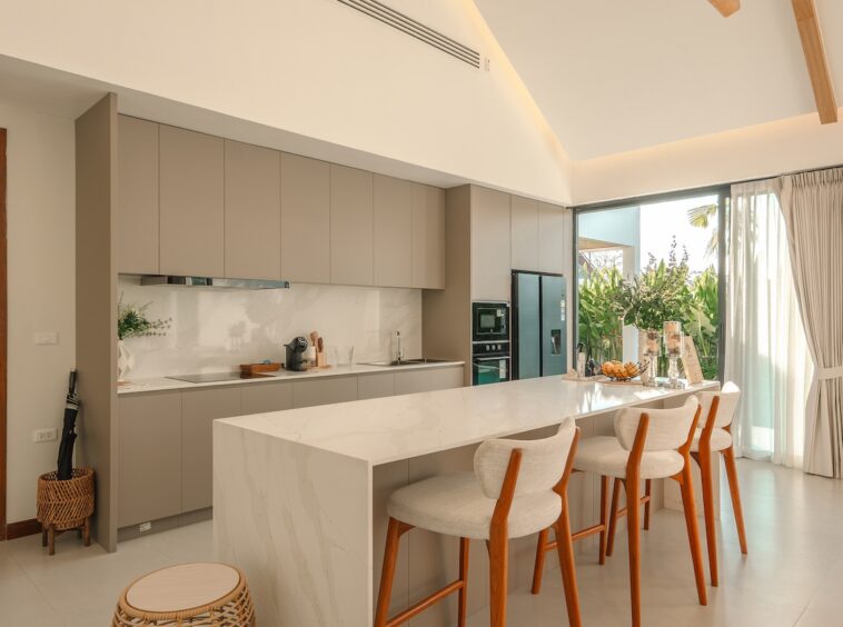 Modern kitchen with a marble island, gray cabinetry, and four wooden-framed bar stools, opening to a sunny patio with plants.