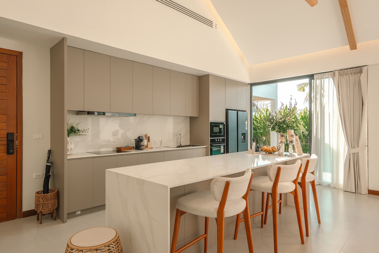 Modern kitchen with a marble island, gray cabinetry, and four wooden-framed bar stools, opening to a sunny patio with plants.