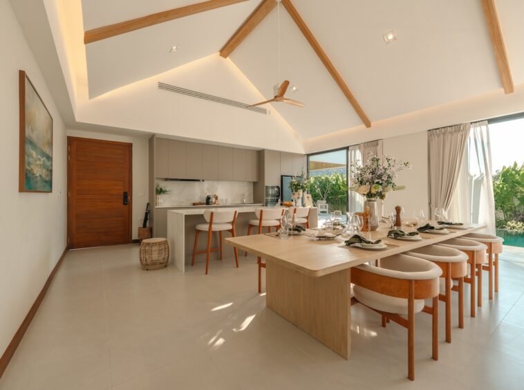 Bright open-plan dining and kitchen area with a vaulted ceiling, wooden beams, and a long dining table set for a meal beside a glass wall overlooking a pool.