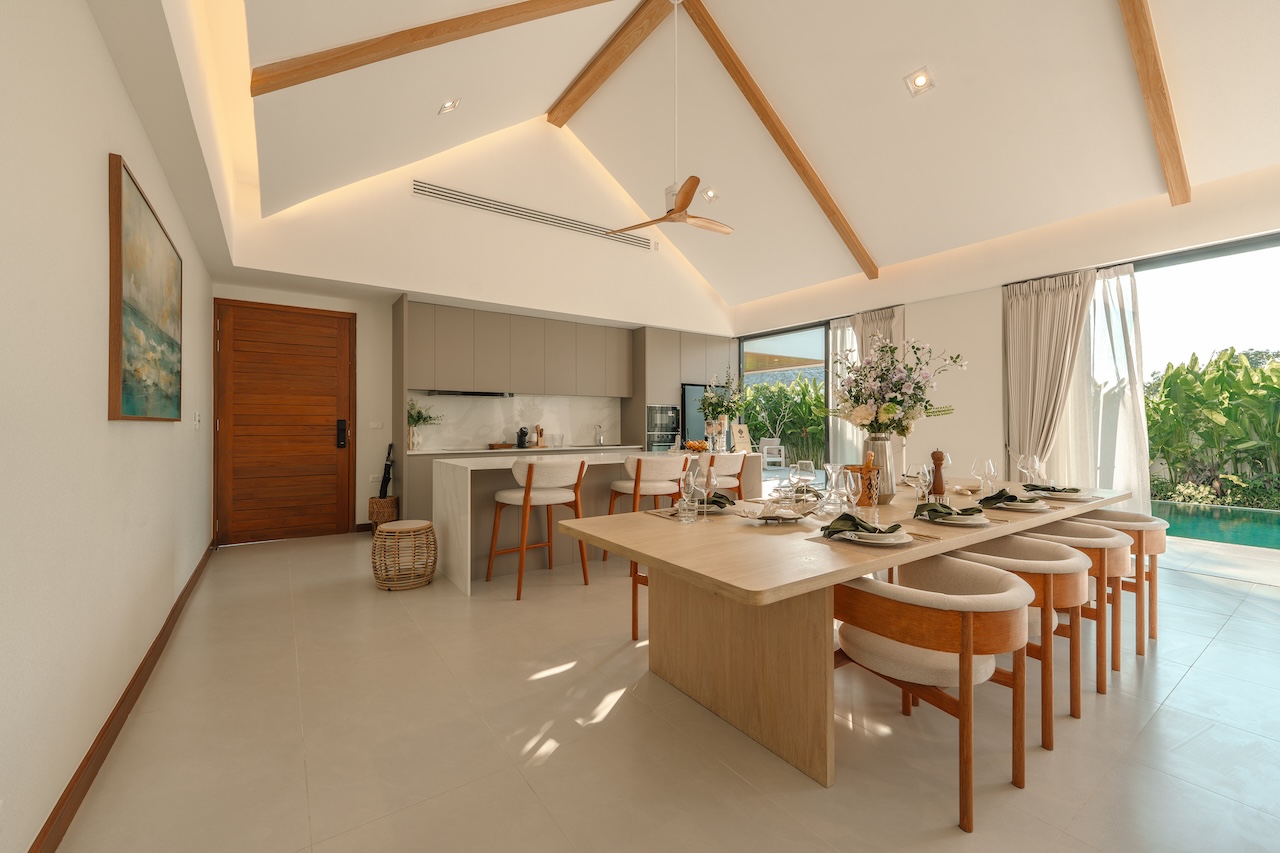 Bright open-plan dining and kitchen area with a vaulted ceiling, wooden beams, and a long dining table set for a meal beside a glass wall overlooking a pool.