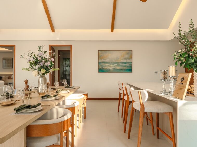 Bright dining room with a long wooden table set for a meal, white upholstered chairs, and floral centerpieces.