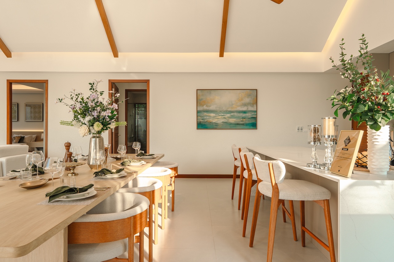 Bright dining room with a long wooden table set for a meal, white upholstered chairs, and floral centerpieces.