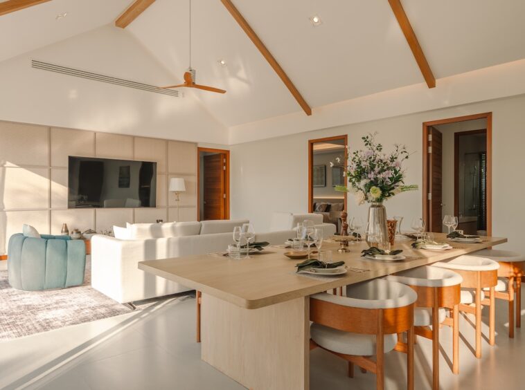 Bright open-plan living and dining room with vaulted ceiling, wooden beams, and a long dining table set for a meal with glassware and a centerpiece.