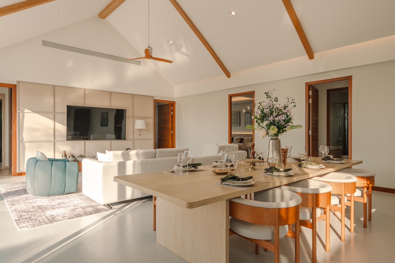 Bright open-plan living and dining room with vaulted ceiling, wooden beams, and a long dining table set for a meal with glassware and a centerpiece.