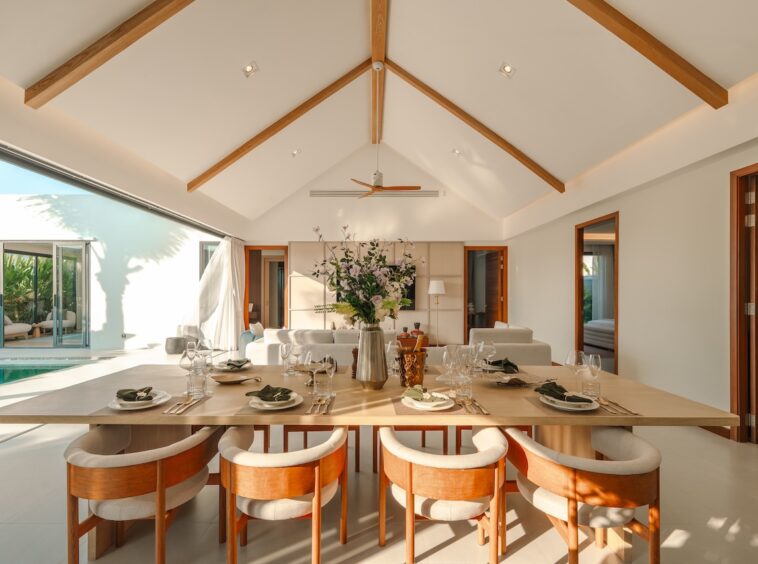 Sunlit dining room with a long wooden table set for eight, cream chairs, and exposed wooden ceiling beams.
