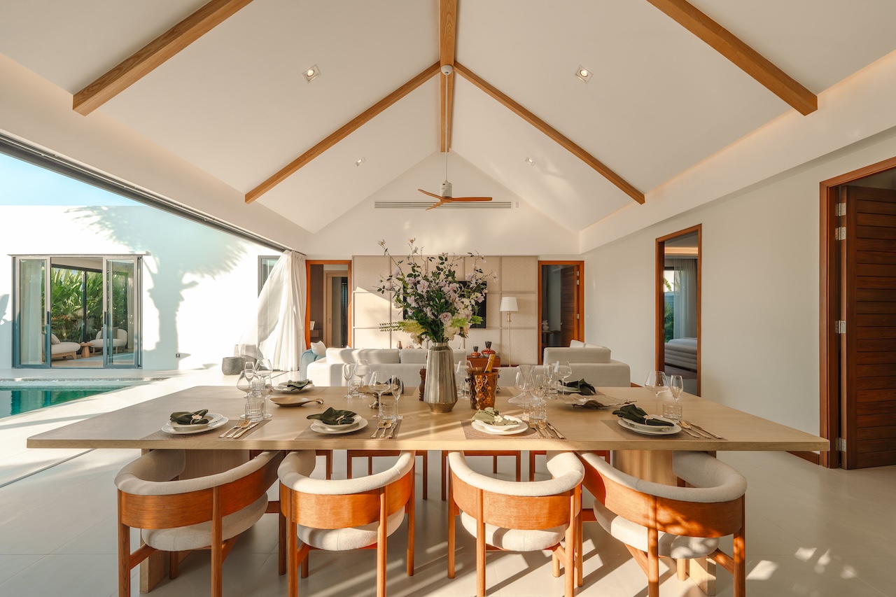 Sunlit dining room with a long wooden table set for eight, cream chairs, and exposed wooden ceiling beams.