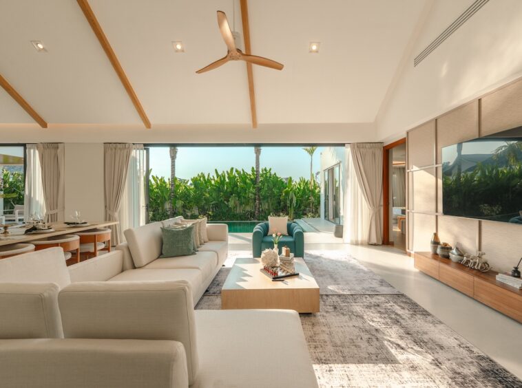 Bright open-plan living room with white sofas, a central coffee table, and a view of a pool and tropical plants outside.