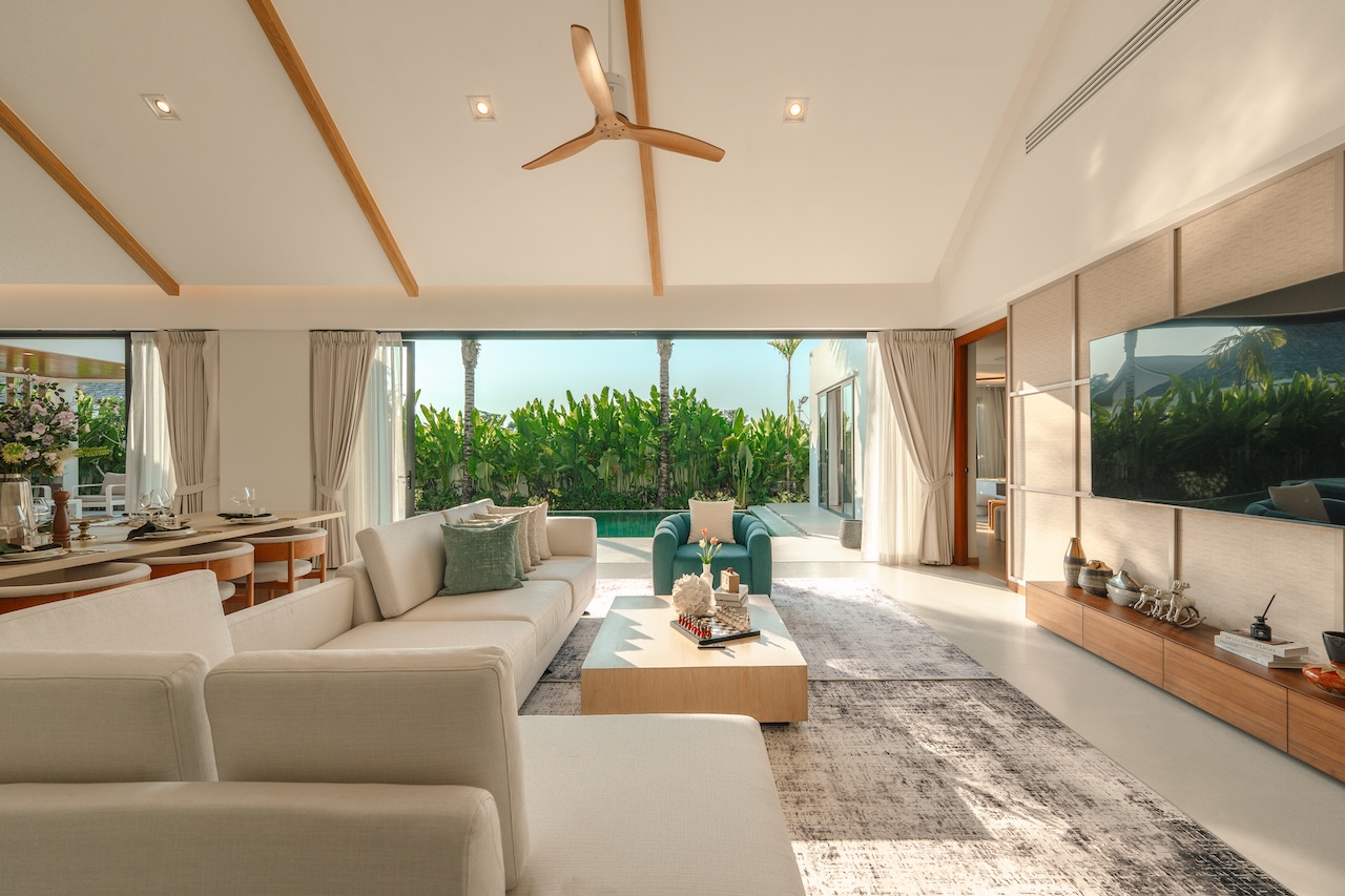 Bright open-plan living room with white sofas, a central coffee table, and a view of a pool and tropical plants outside.