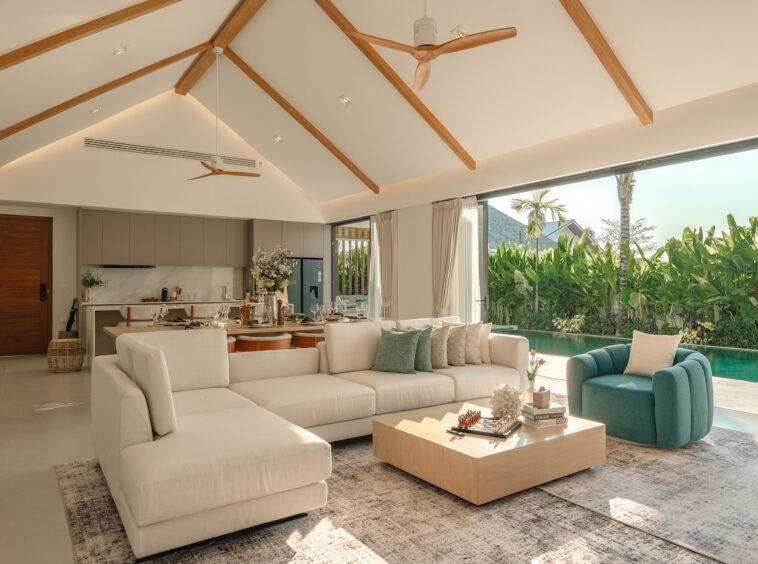 Bright open-plan living room with white sectional sofa, teal armchair, and a low wooden coffee table beside a poolside view.