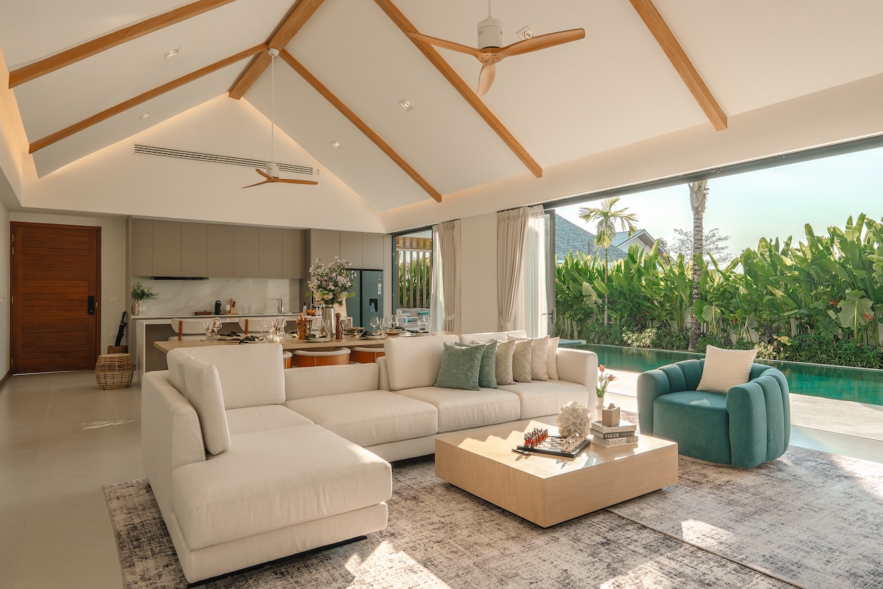 Bright open-plan living room with white sectional sofa, teal armchair, and a low wooden coffee table beside a poolside view.