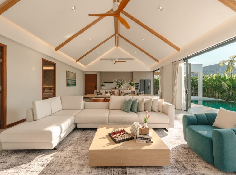 Open-concept living room with a white sectional, teal chair, and a low wooden coffee table, opening to a pool area through large glass doors.