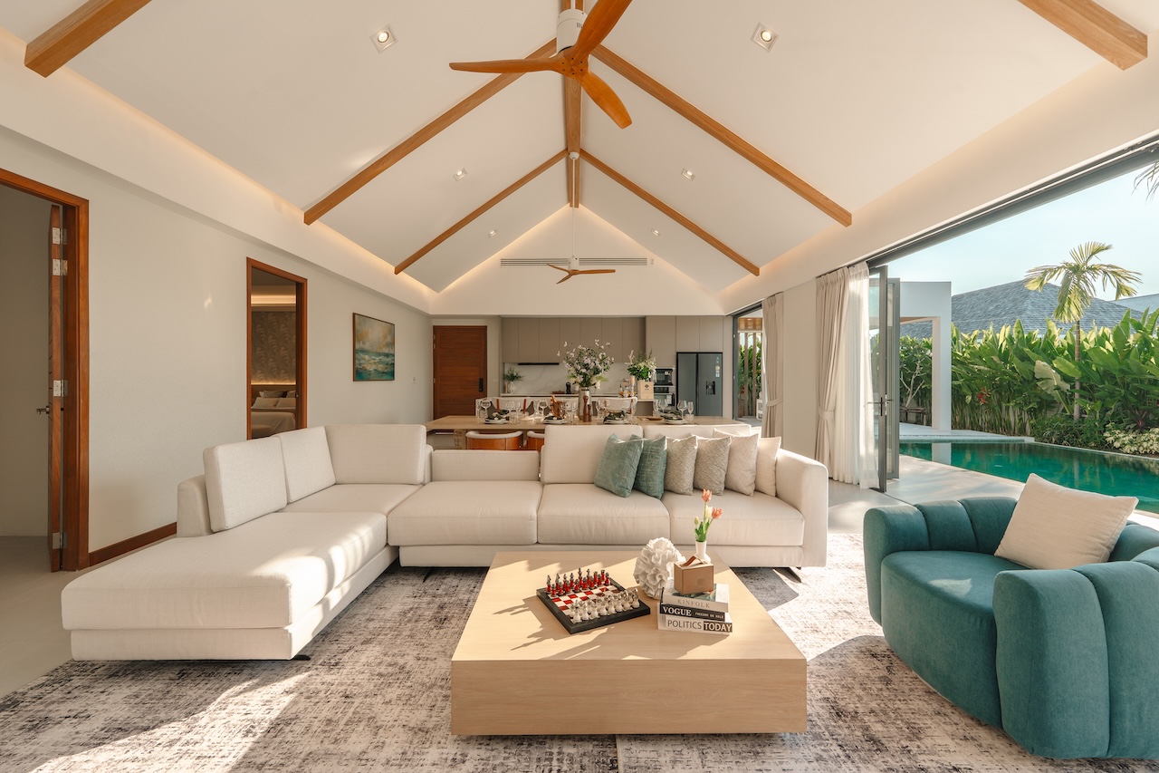 Open-concept living room with a white sectional, teal chair, and a low wooden coffee table, opening to a pool area through large glass doors.