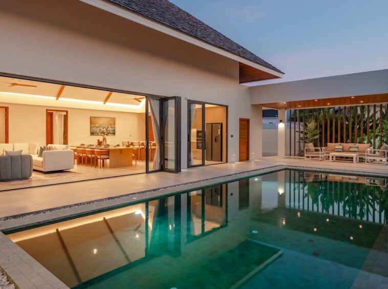 Modern villa with an open-plan living/dining area and a rectangular pool along the terrace at dusk.