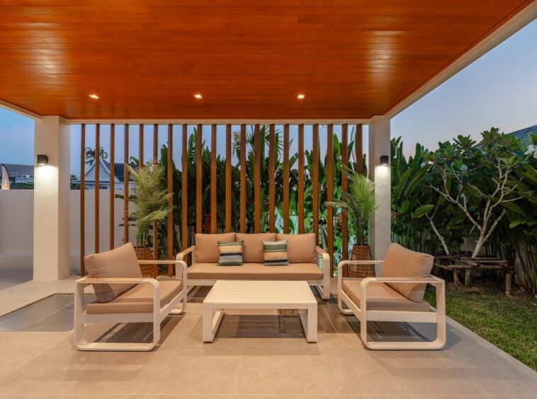 Outdoor lounge area with beige cushioned sofa and two matching chairs under a wooden ceiling and slatted backdrop, tropical plants nearby.
