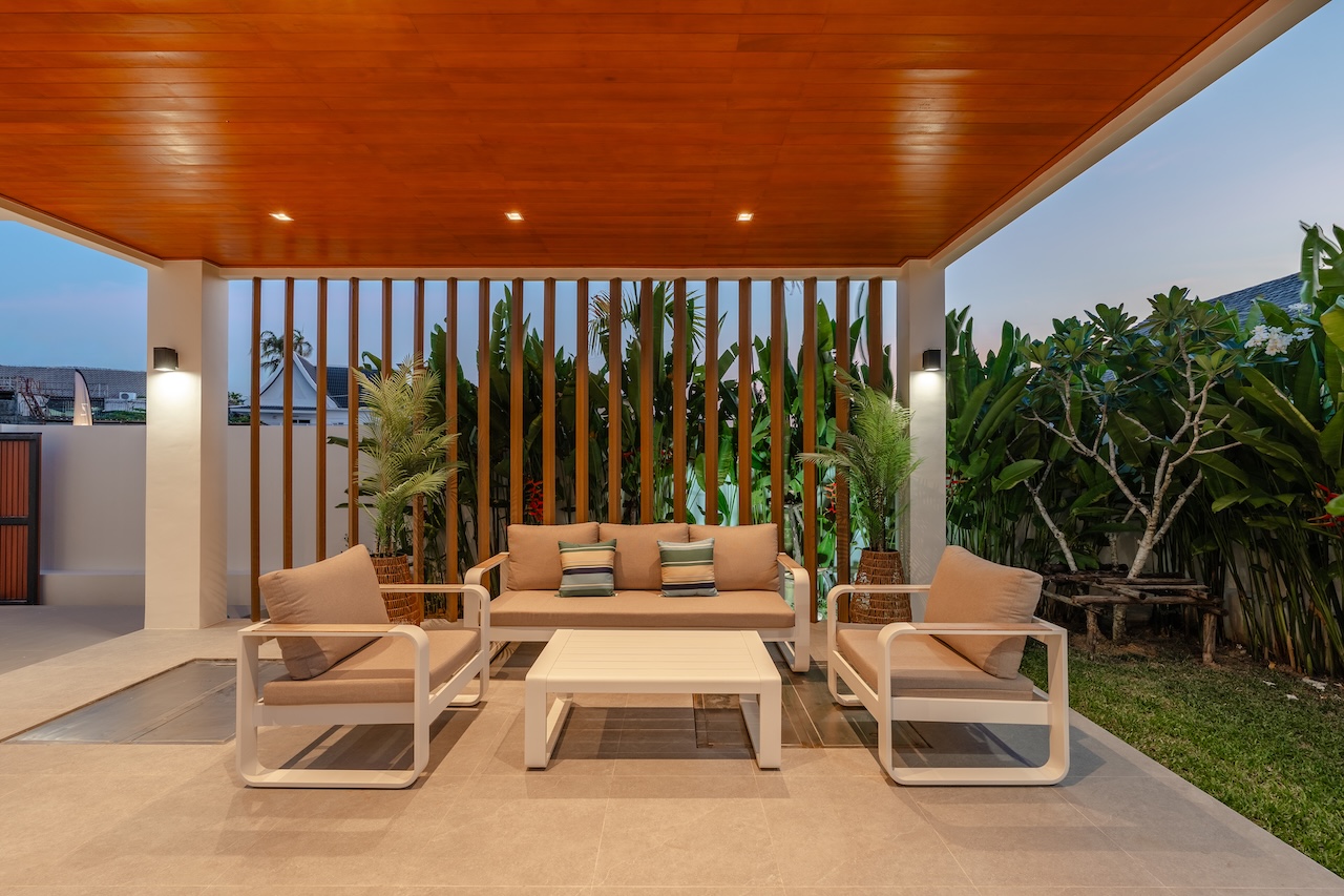 Outdoor lounge area with beige cushioned sofa and two matching chairs under a wooden ceiling and slatted backdrop, tropical plants nearby.