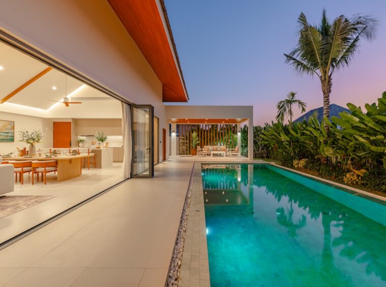 Open-plan villa with indoor dining area and kitchen, sliding glass doors opening to a turquoise pool at dusk, tropical garden beyond.