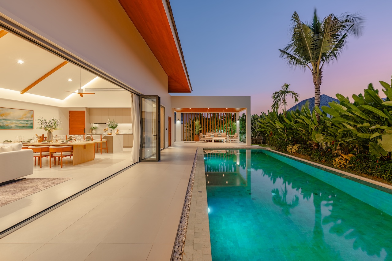 Open-plan villa with indoor dining area and kitchen, sliding glass doors opening to a turquoise pool at dusk, tropical garden beyond.