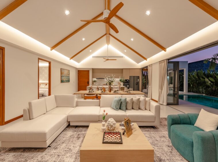 Open-concept living area with a cream sectional sofa, teal chair, wooden coffee table, and a dining table in the background, near a pool.