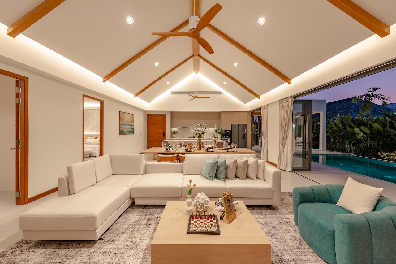 Open-concept living area with a cream sectional sofa, teal chair, wooden coffee table, and a dining table in the background, near a pool.