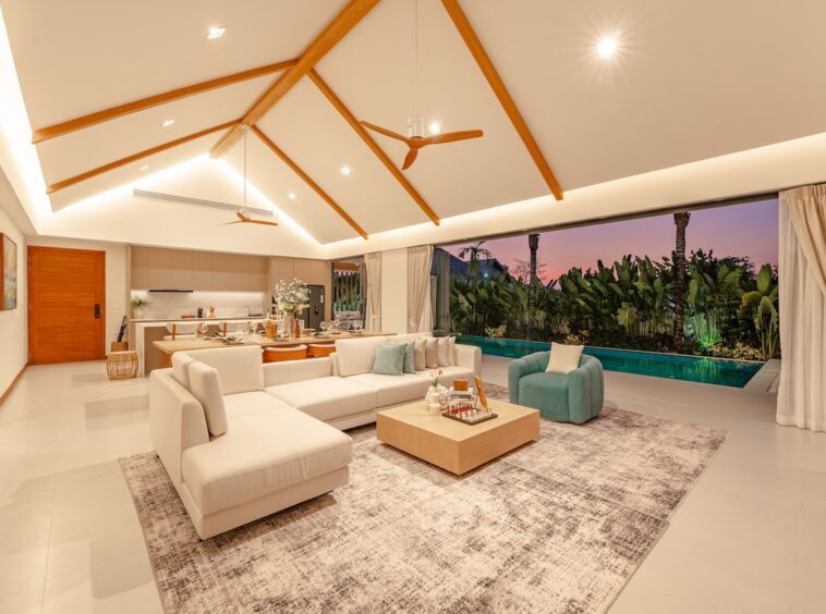 Open-plan living room with white sectional sofa, wooden coffee table, and turquoise chairs facing a pool at sunset through large windows. A high vaulted ceiling with wooden beams dominates the space.