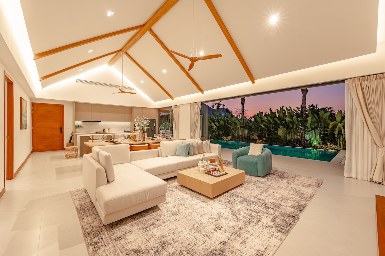 Open-plan living room with white sectional sofa, wooden coffee table, and turquoise chairs facing a pool at sunset through large windows. A high vaulted ceiling with wooden beams dominates the space.