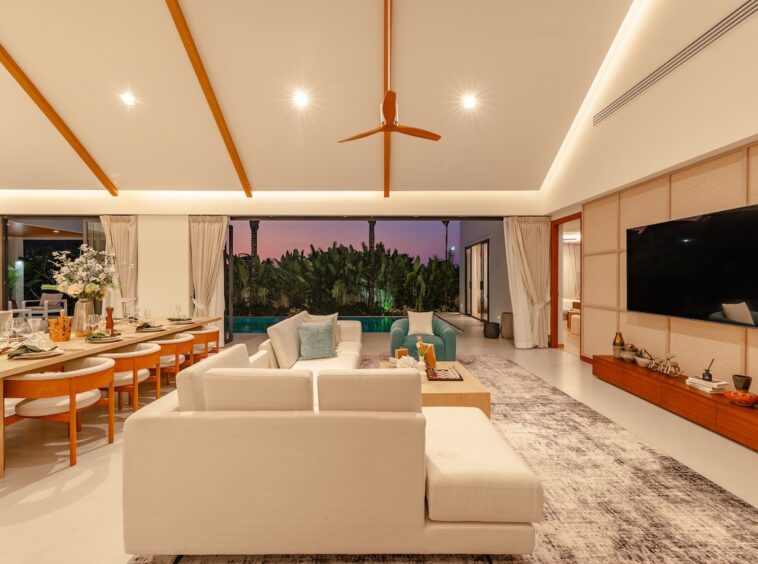 Open-plan living and dining area at dusk, with white sofas, a long wooden dining table, and sliding doors to a tropical garden outside.