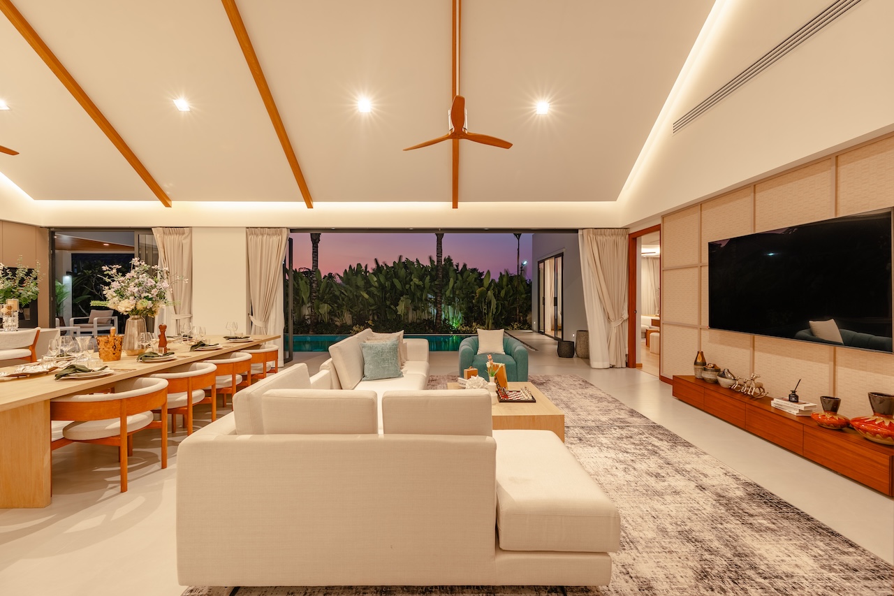 Open-plan living and dining area at dusk, with white sofas, a long wooden dining table, and sliding doors to a tropical garden outside.
