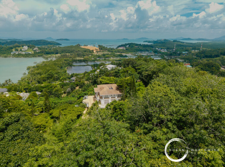 Aerial view of a lush, hilly landscape with a large house in the foreground, terraced ponds, and the sea on the horizon.