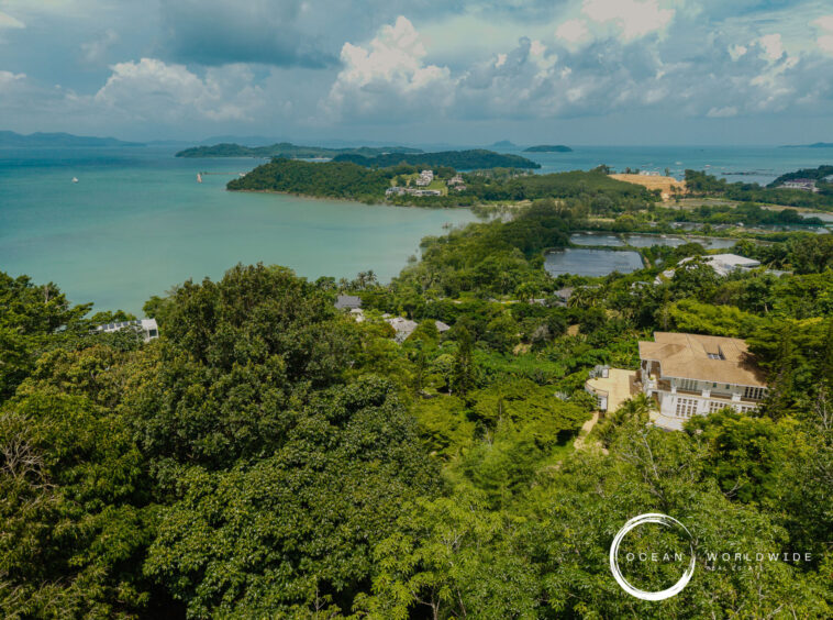 A tropical coastal landscape with lush green foreground, turquoise water, and small islands under a cloudy sky as seen from above.