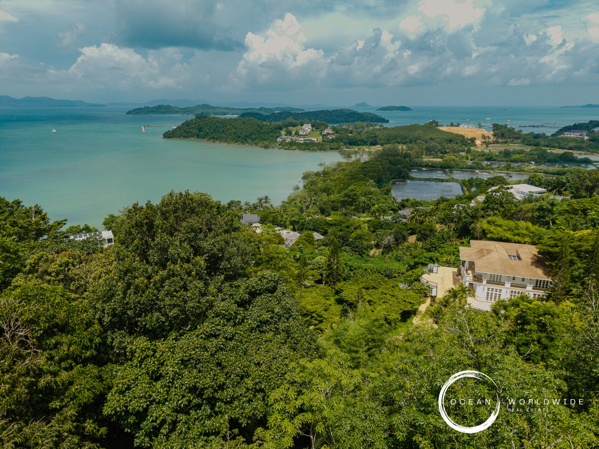 A tropical coastal landscape with lush green foreground, turquoise water, and small islands under a cloudy sky as seen from above.