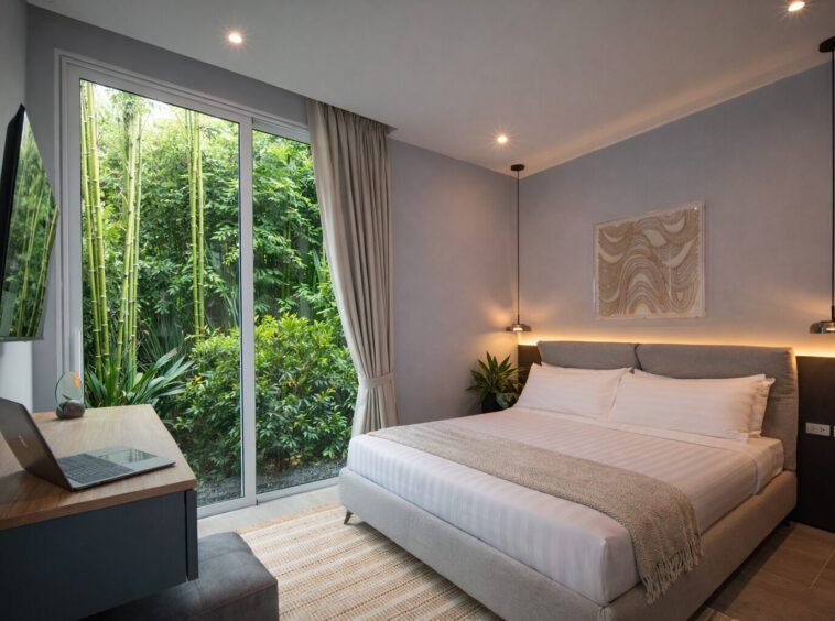 Hotel bedroom with a large bed, pendant lights, and a desk facing a glass sliding door to a green garden outside.
