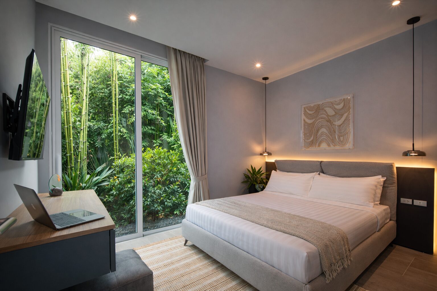 Hotel bedroom with a large bed, pendant lights, and a desk facing a glass sliding door to a green garden outside.