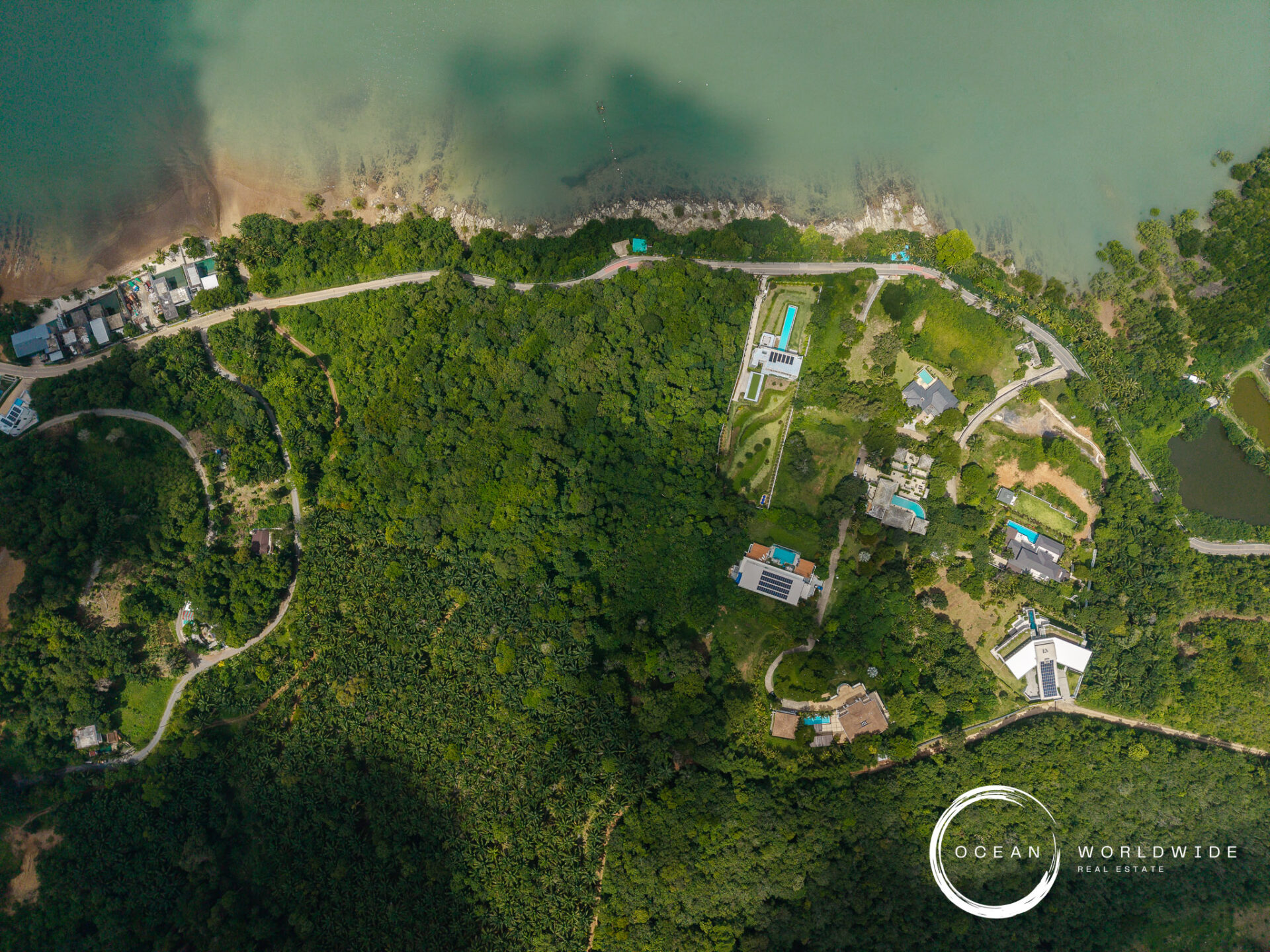 Aerial view of a coastal resort area with dense forest, a road running along the shore, and scattered buildings and pools; Ocean Worldwide logo bottom-right.