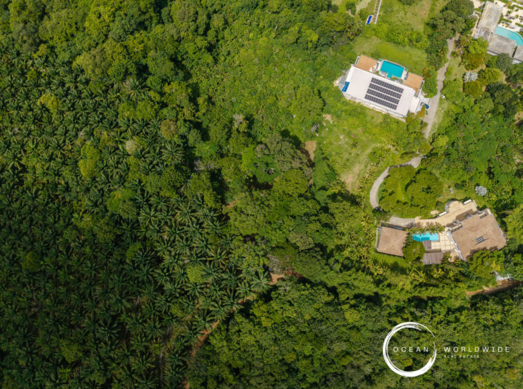 Aerial view of dense tropical forest with a modern house, pool, and manicured lawn on the right; Ocean Worldwide Real Estate watermark bottom-right.
