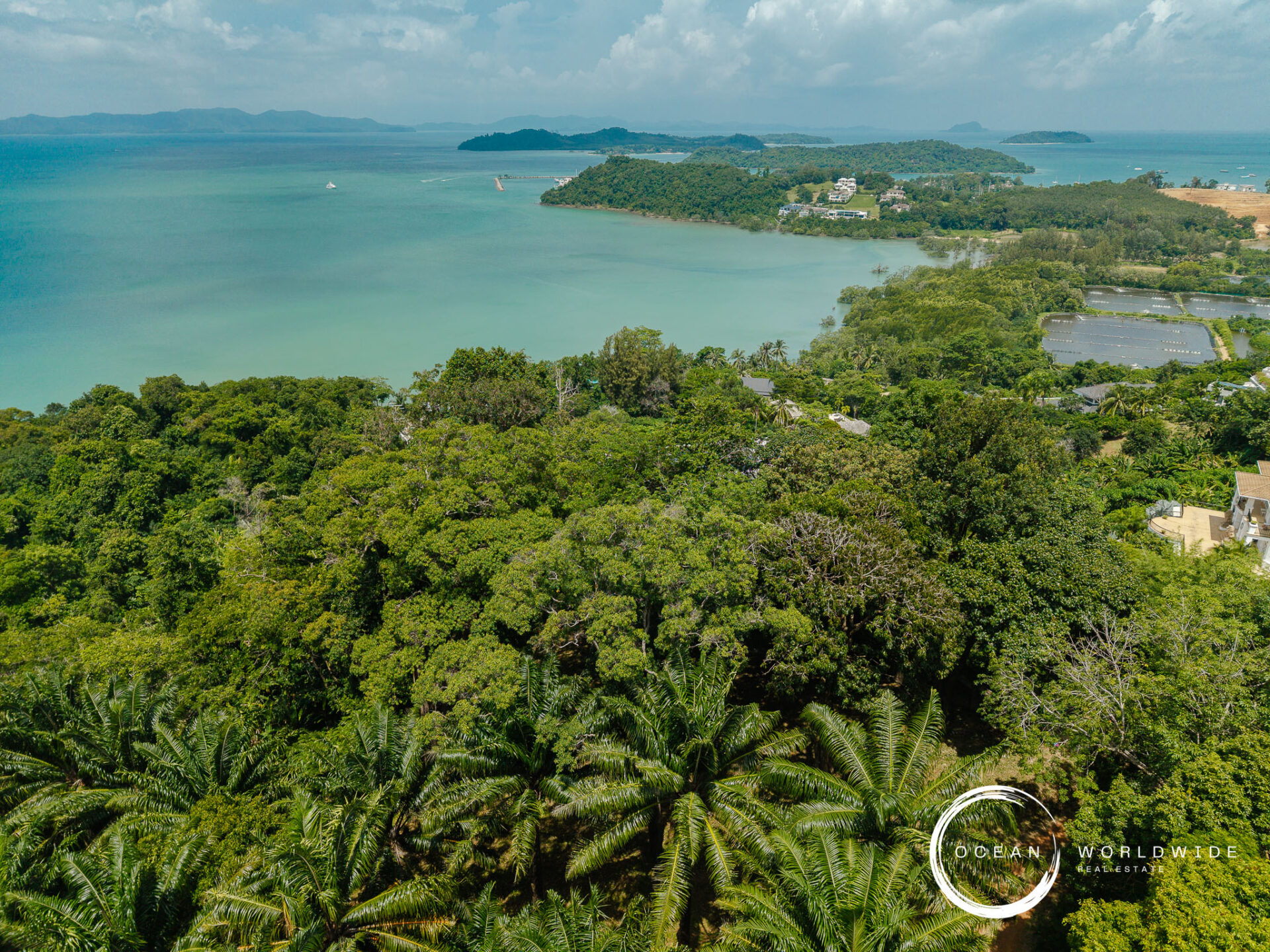 Aerial view of dense tropical forest along a turquoise coastline with small islands in the distance and a few buildings visible; Ocean Worldwide logo bottom right.