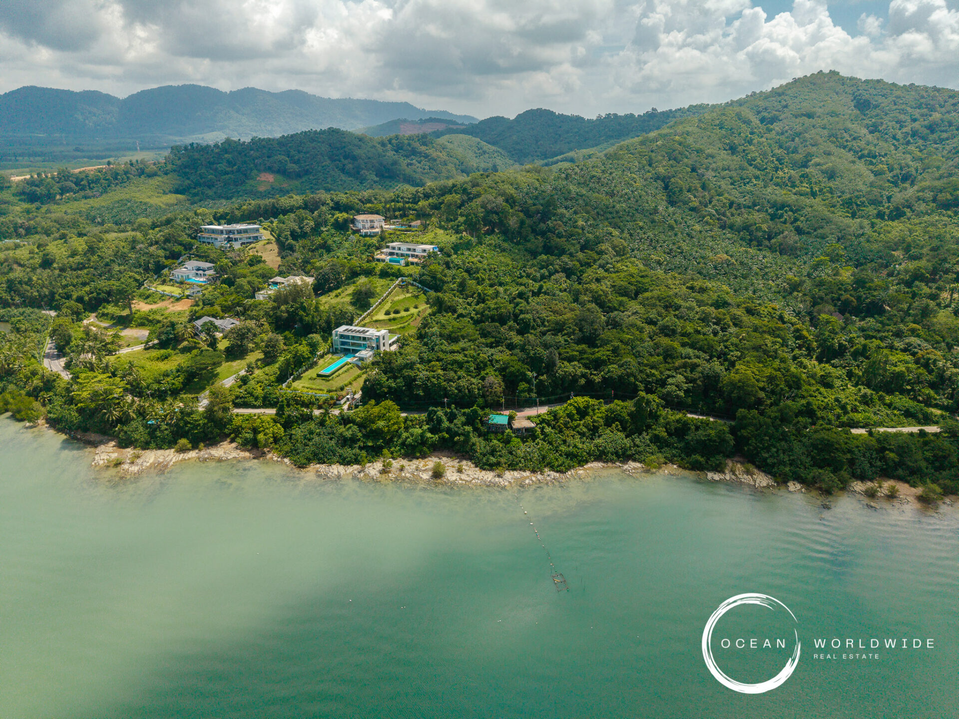 Aerial view of a green hillside coastline with scattered houses and a pool near the shore; Ocean Worldwide Real Estate logo bottom-right.