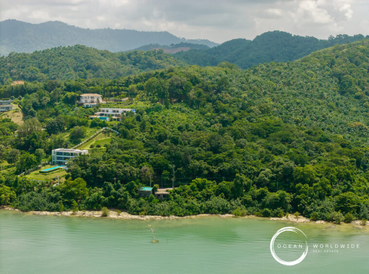 Lush hillside with modern houses along a tropical coastline, dense trees by the water, and a watermark reading 'Ocean Worldwide Real Estate' bottom right.