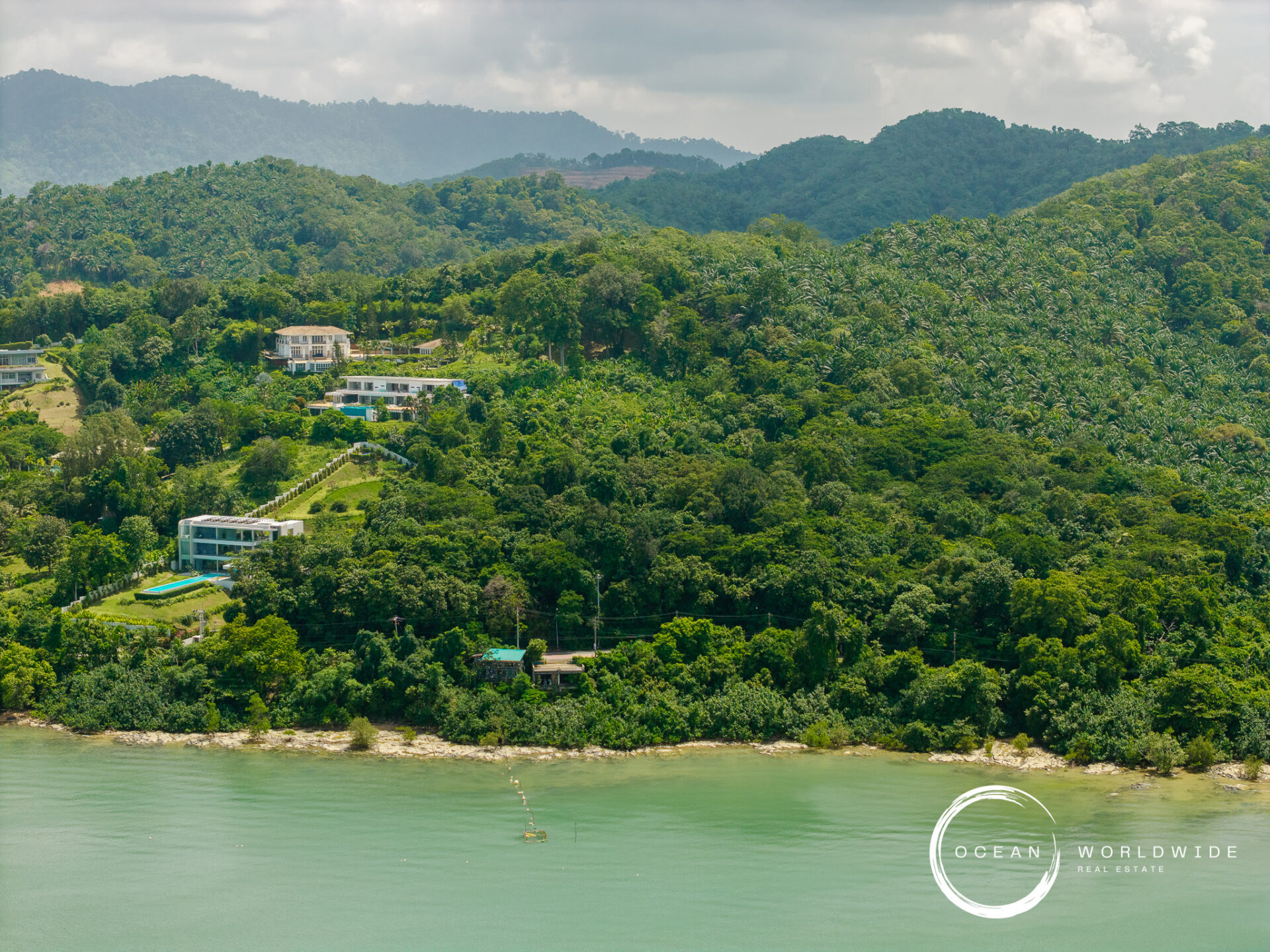 Lush hillside with modern houses along a tropical coastline, dense trees by the water, and a watermark reading 'Ocean Worldwide Real Estate' bottom right.