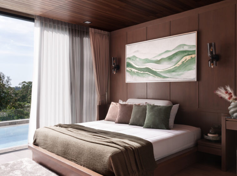 Modern bedroom with dark wood paneled walls, king bed and green/brown throw pillows, large landscape painting, and bedside sconces; sliding doors to a balcony overlook a pool and trees.