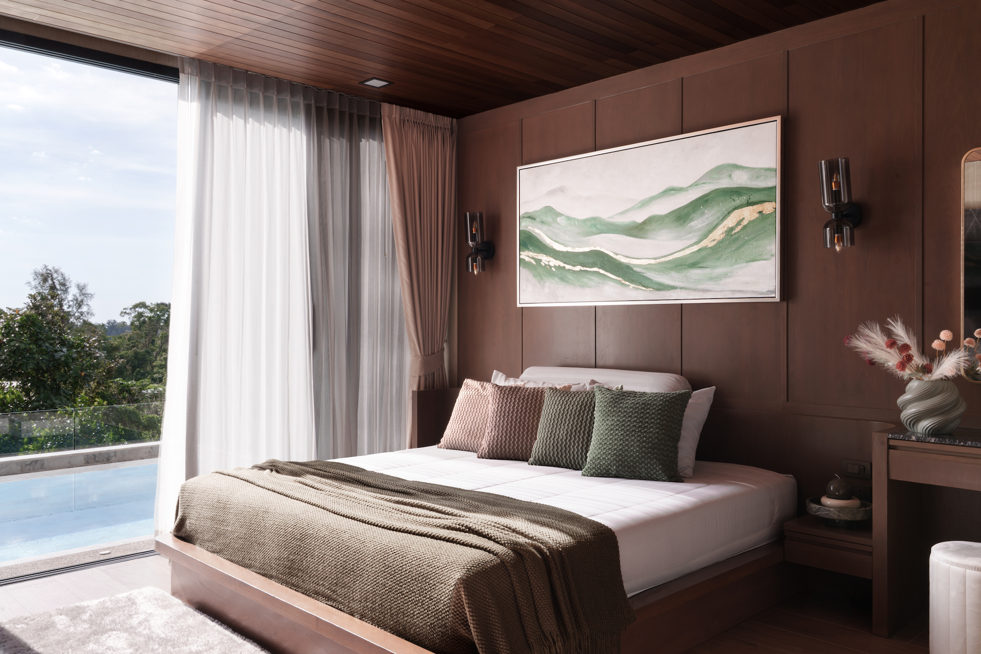 Modern bedroom with dark wood paneled walls, king bed and green/brown throw pillows, large landscape painting, and bedside sconces; sliding doors to a balcony overlook a pool and trees.