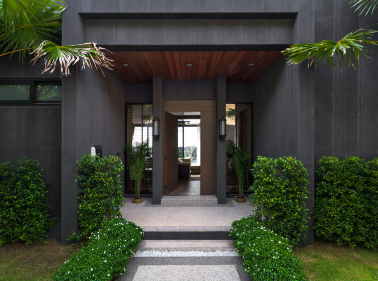 Modern house entrance with dark gray exterior, open wooden doorway, and symmetrical plants framing a pebble path to the door.