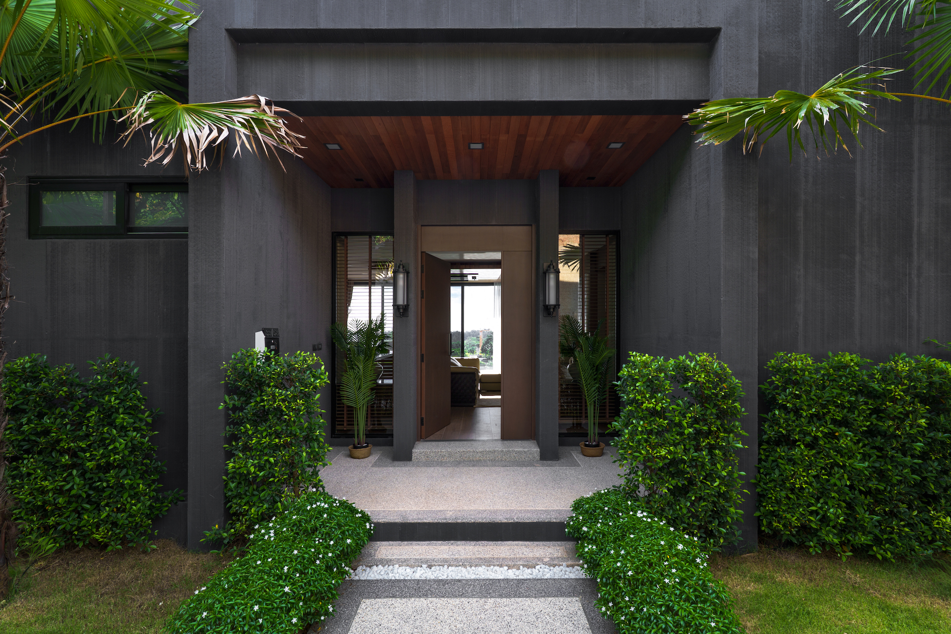 Modern house entrance with dark gray exterior, open wooden doorway, and symmetrical plants framing a pebble path to the door.