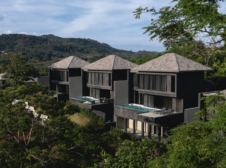 Row of modern black villas with pyramid roofs and glass balconies beside a swimming pool, set against green hills and trees.