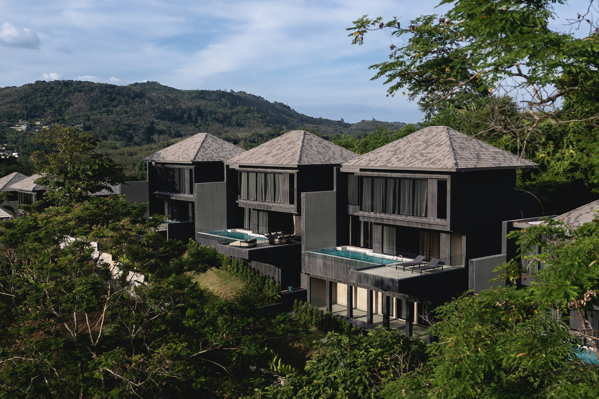 Row of modern black villas with pyramid roofs and glass balconies beside a swimming pool, set against green hills and trees.