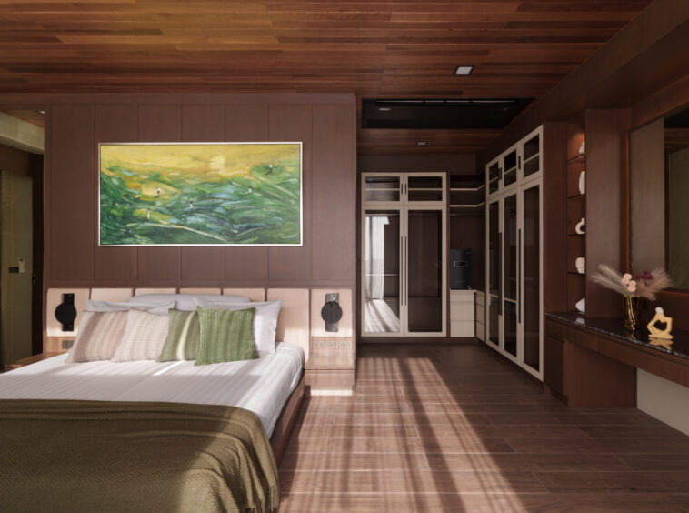 Bedroom with dark wood paneling, a large white bed with green beige cushions, and a vibrant landscape painting above. Shaded sunlight across the floor.