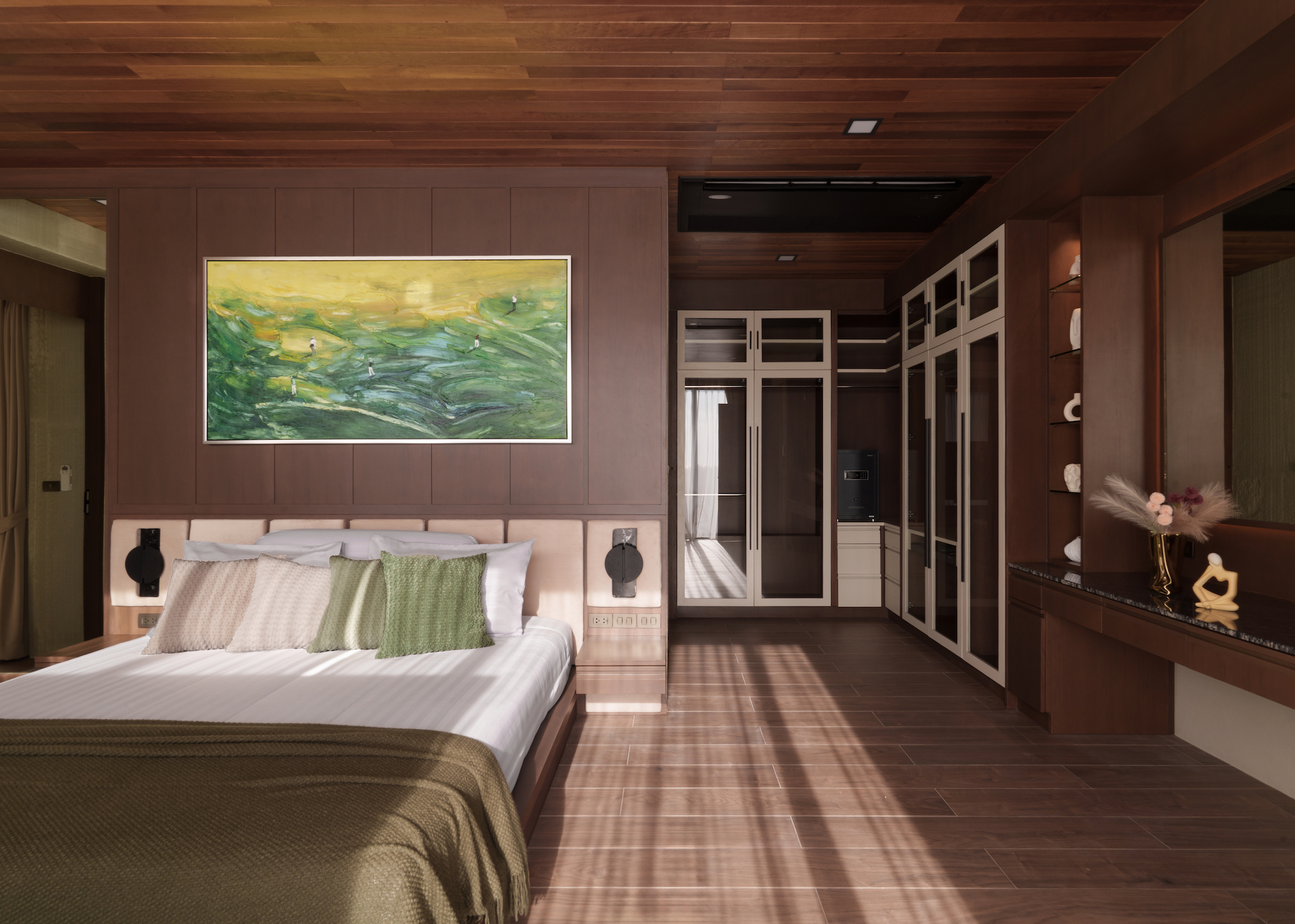 Bedroom with dark wood paneling, a large white bed with green beige cushions, and a vibrant landscape painting above. Shaded sunlight across the floor.